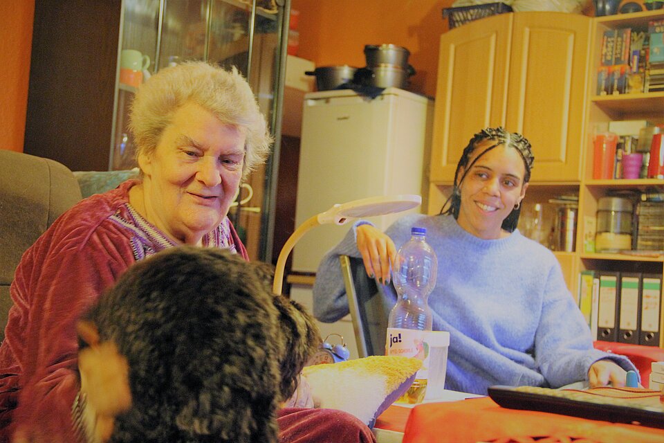 Eine alte Frau und eine junge Frau sitzen lachend nebeneinander am Tisch, die alte Frau streichelt einen Hund