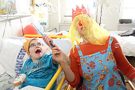 Ein Junge liegt in einem Krankenbett, eine Frau im Clownskostüm sitzt neben ihm und lässt Seifenblasen aufsteigen, beide freuen sich.