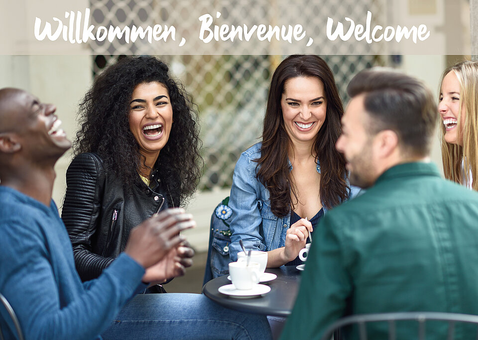 Vier Personen sitzen am Tisch und lachen, darüber Text: Willkommen, Bienvenue, Welcome