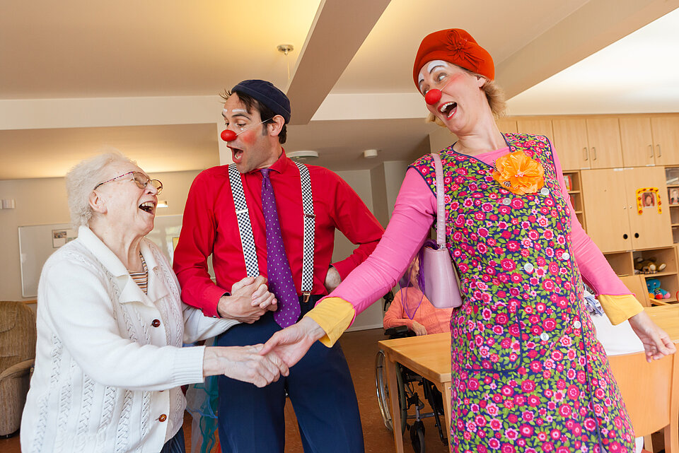 Zwei Clowns halten die Hände einer Seniorin, alle Drei lachen.