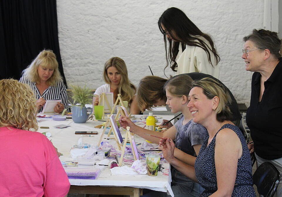 Viele Frauen und Mädchen unterschiedlichen Alters sitzen an kleinen Staffeleien um einen Tisch und malen, einige schauen konzentriert, ander lachen. 