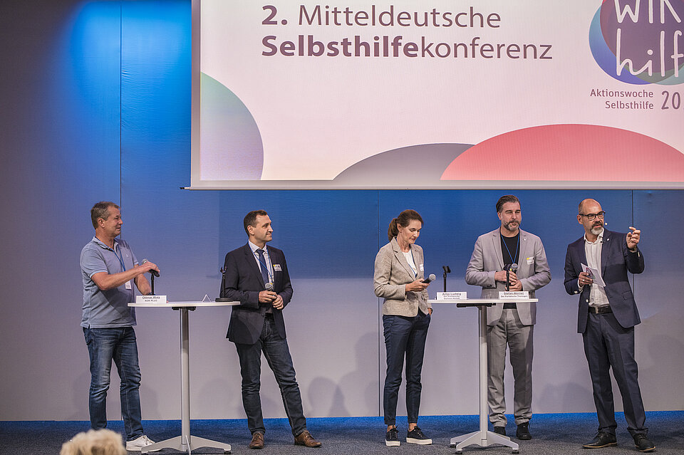 Vier Männer und eine Frau stehen auf einer Bühne mit Mikrofonen an Tischen, im Hintergrund steht: 2. Mitteldeutsche Selbsthilfekonferenz. 