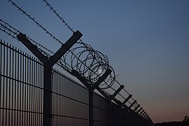 In der Abenddämmerung ist ein Zaun zu sehen, der nach oben hin mit Stacheldraht begrenzt ist, dahinter scheint der Mond.