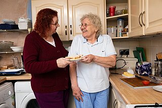 Eine ältere und eine jüngere Frau stehen zusammen in einer Küche und lächeln sich an