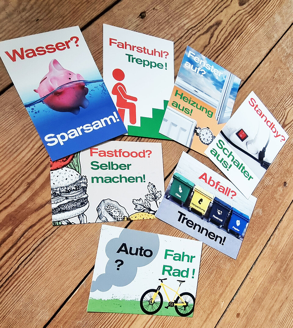 Auf einem Holzboden liegen Postkarten mit Energiespartipps wie "Wasser? Sparen", "Fenster auf? Heizung aus!", "Auto? Fahrrad!"