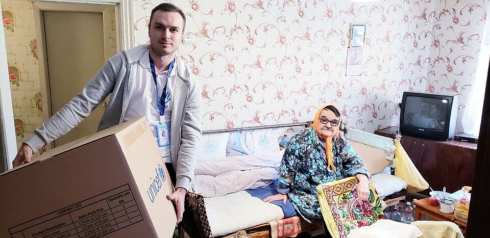 Ein junger Mann trägt eine große Pappkiste in die Wohnung einer alten Frau, die auf einem Sofa sitzt.