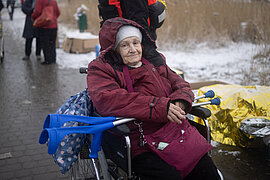 Eine alte Frau sitzt müde lächelnd in einem Rollstuhl, auf ihrem Rollstuhl liegen Gehhilfen, im Hintergrund liegt Schnee.