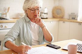 Eine ältere Dame mit weißen Haaren sitzt konzentriert an ihrem Küchentisch und berechnet etwas. Neben ihr liegt ein Taschenrechner.