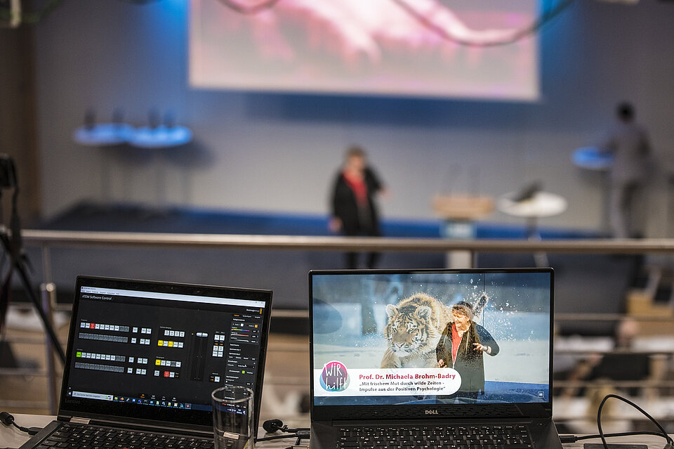Im Hintergrund: Eine Frau steht auf einer Bühne mit einem Mikrophon. Im Vordergrund: Die Frage ist noch einmal auf einem Laptopbildschirm zu sehen mit dem Foto eines Tigers.