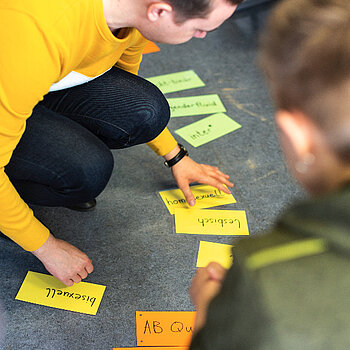 Eine Teamer_in legt Begriffkarten auf den Boden in die Mitte eines Stuhlkreises. Bild: MikaWisskirchen