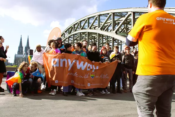 Eine Gruppe junger Menschen steht zusammen hinter einem Transparent auf dem "anyway" steht, im Hintergrund der Kölner Dom.