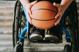 Rollstuhlfahrende Person hält Basketball auf Fußhöhe. Nahaufnahme von vorn