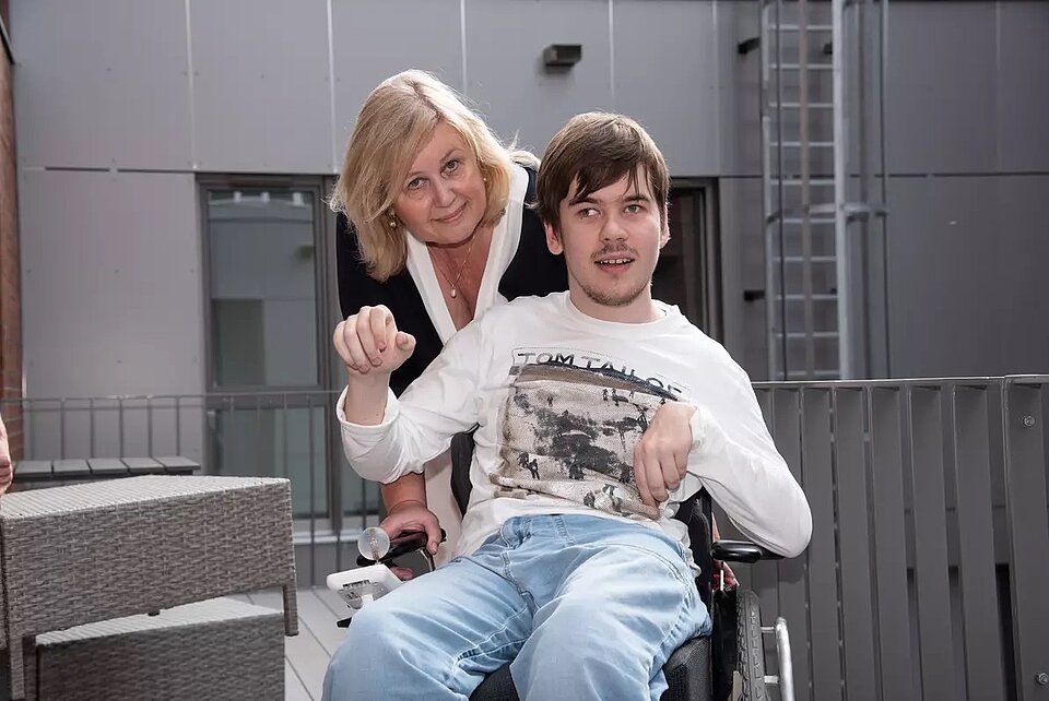 Vor einem Neubau steht eine Frau und vor ihr ein junger Mann im Rollstuhl. Es ist Ingrid Bolz mit ihrem Sohn Andreas.