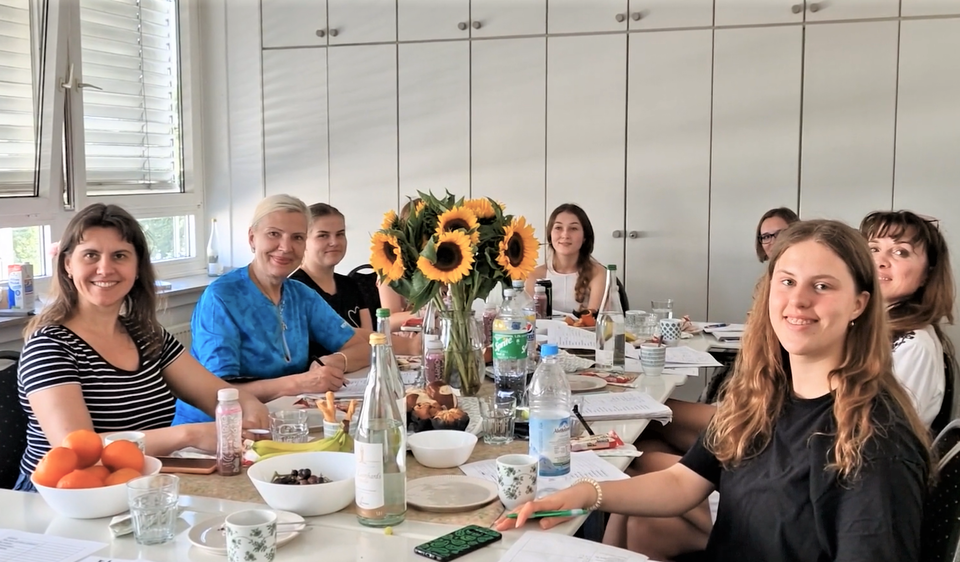 Frauen verschiedenen Alters sitzen mit Schreibuntensilien lachend an einem Tisch zusammen, auf dem ein großer Strauß Sonnenblumen steht. 