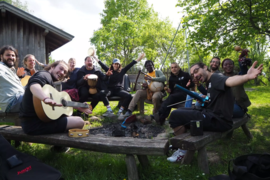 Viele unterschiedliche Menschen sitzen im Sonnenschein fröhlich im Kreis mit Musikinstrumenten an einer Lagerfeuerstelle.