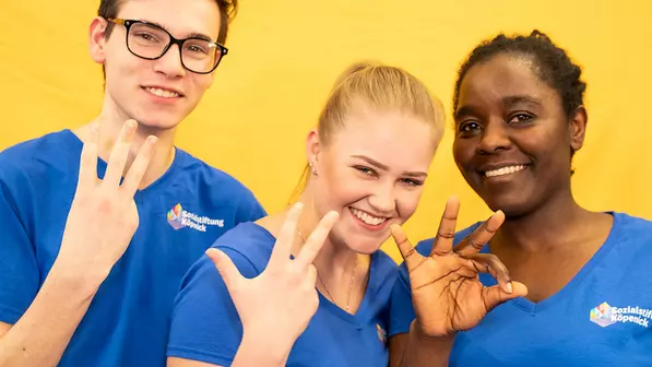 Drei junge Menschen unterschiedlicher Hautfarbe zeigen auf drei unterschiedlichen Wegen mit ihren Fingern die Zahl 3 und lachen. Auf ihren Pflege-Shirts steht: Sozialstiftung Köpenick.