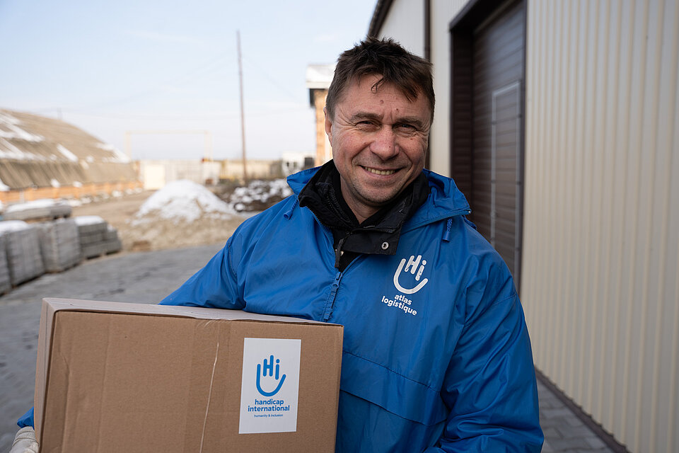 Ein Mann trägt einen großen Karton, er trägt eine Jacke mit dem Logo von Handicap International und lächelt.