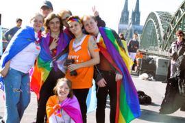 Eine Gruppe lachender Jugendlicher mit Regenbogenfahnen steht zusammen, im Hintergrund der Kölner Dom