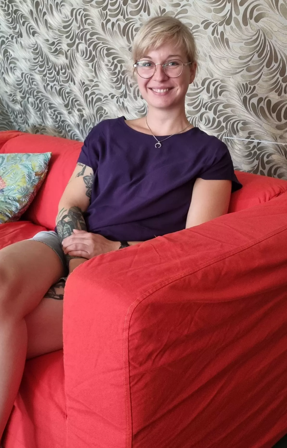 Eine junge Frau mit blonden Haaren, Brille und Tattoos auf dem Arm sitzt lächelnd auf einer Couch von einer geblümten Tapete