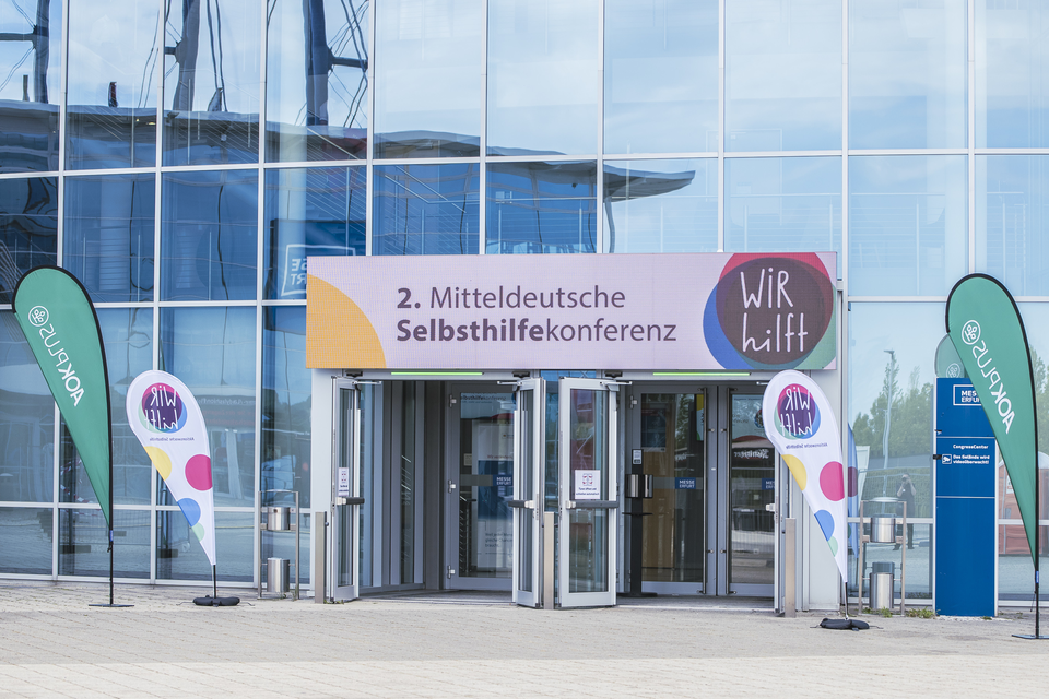 Vor einer Eingangshalle ist ein bunter Banner zu sehen: 2. Mitteldeutsche Selbsthilfekonferenz