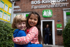 Zwei Kinder umarmen sich und lachen, im Hintergrund das Logo von SOS Kinderdorf