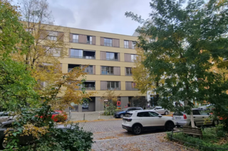 Ein vierstöckiges Wohngebäude mit Balkonen vor einer Straße umrahmt von Bäumen.