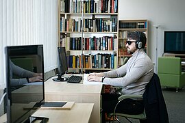 Eine männliche Person sitzt in einer Bibliothek und liest in einem Buch mit Brailleschrift. Er hat Kopfhörer auf und trägt eine Sonnenbrille. Vor ihm steht ein PC. (Symbolbild)