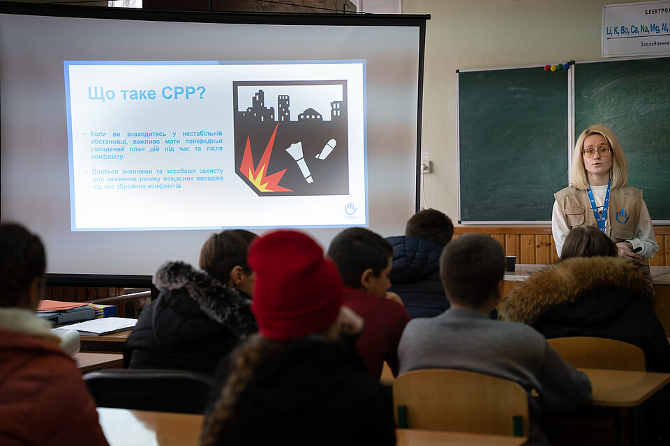 Eine Frau spricht vor einer Gruppe von Menschen, die in Reihen auf Stühlen sitzen, auf einem an eine Wand projezierten Bild erkennt man eine Rakete. 