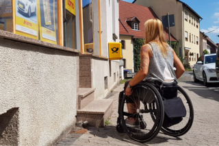 Rollstuhlfahrerin steht auf einer Treppe, die zur Poststelle führt.