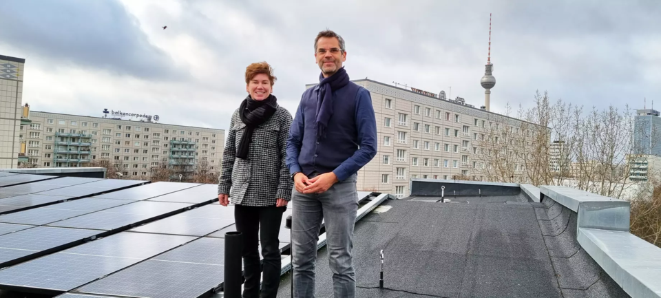 Ein Mann und eine Frau stehen auf einem Dach, auf welchem Solarplatten liegen, dahinter ist der Berliner Fernsehturn zu sehen. 