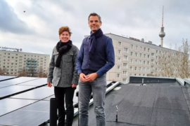 Ein Mann und eine Frau stehen auf einem Dach, auf welchem Solarplatten liegen, dahinter ist der Berliner Fernsehturn zu sehen. 