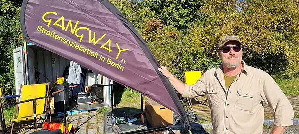 Ein Mann mittleren Altern in Sonnenbrille steht mit einem Grinsen an einer Fahne, auf welcher steht: "Gangway. Straßensozialarbeit in Berlin."