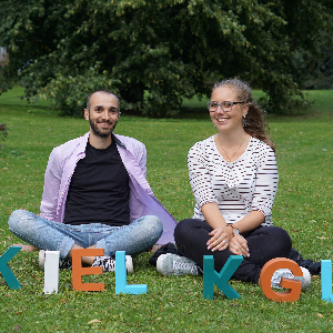 Tandem von kulturgrenzenlos. Junger Mann mit Fluchthintergrund und junge Frau sitzen auf einer Wiese und schauen in die Kamera, sie lächeln.