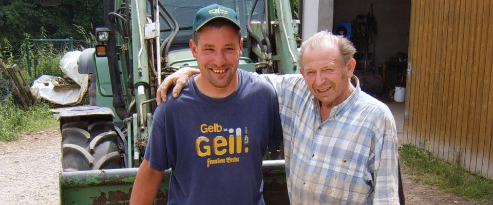 Zwei Menschen, einer davon jung, der andere älter, stehen in Arbeitskleidung vor einem Traktor. Bild mit freundlicher Genehming von integra menshc, Bamberg