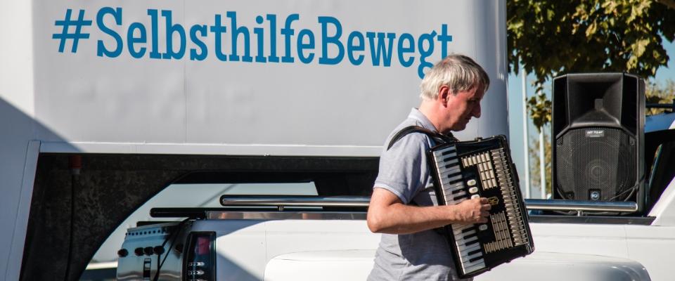 Mann mit Akkordeon vor einem Truck mit der Aufschrift "Selbsthilfe bewegt"