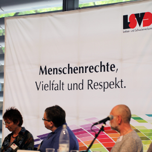 Foto mit einem Podium vom Verbandstag. Im Hintergrund unser Motto „Menschenrechte, Vielfalt und Respekt“