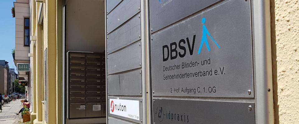 Straßenansicht der Rungestraße in Berlin. An der Fassade des Vorderhauses hängt das Schild der DBSV-Geschäftsstelle in Berlin. Es zeigt die Illustration eines Stockgängers von der Seite in Cyan-Blau, links daneben in Versalien "DBSV".