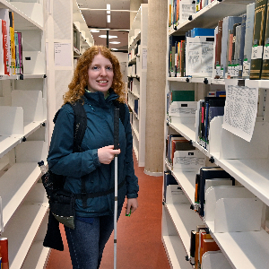 Eine Studentin mit weißem Stock steht in der Universitätsbibliothek zwischen zwei Bücherregalen
